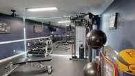 291 North Gym is a thoughtfully designed, fully equipped fitness space that delivers a professional gym experience in a compact, efficient layout. The room is finished with bold navy blue walls accented by a crisp white chair rail, creating a sharp, motivating atmosphere complemented by bright overhead fluorescent lighting and ceiling fans for ventilation and comfort. Durable speckled rubber flooring covers the entire space, providing cushioning and traction ideal for high-intensity training. The cardio zone features multiple treadmills, an elliptical trainer, and a stationary bike, each positioned in front of wall-mounted flat-screen TVs for an engaging workout experience. A large floor-to-ceiling mirror along one wall visually expands the space and aids in form monitoring. The strength training area is well-appointed with a multi-function cable machine, a full dumbbell rack ranging across various weights, adjustable benches, stability balls, resistance bands, and medicine balls. A branded "291 North Gym" sign serves as a focal point, adding a personalized identity to the space. Workout reference charts are posted on the walls for added utility. This gym is designed to support a wide range of fitness goals, from cardio conditioning to strength and functional training.