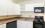 This single-floor unit offers a clean, functional layout designed for comfortable everyday living. Rich hardwood flooring flows consistently throughout the space, lending warmth and visual continuity to each area.

The kitchen serves as a central highlight, featuring crisp white cabinetry that extends to the ceiling for ample storage, stone-look laminate countertops, and a full suite of black appliances including a refrigerator, electric range with overhead microwave, and dishwasher. A peninsula island with matching countertop provides additional prep space and casual dining potential, while a pendant light above adds a finishing touch.

The living area is open and bright, with a front-facing window that draws in natural light and a wall-mounted HVAC unit for climate control. The space is generously proportioned and ready to accommodate a variety of furniture configurations.

The bathroom is neatly finished with white cabinetry, stone-look countertops, hardwood flooring, and a full tub-and-shower combination with a curved curtain rod.

A dedicated utility room houses a stacked washer and dryer unit alongside a water heater, with wire shelving for additional storage — a practical convenience within the unit itself. An additional room provides versatile space suitable for use as a bedroom or flex room.