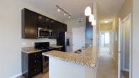 Located at 1444 Chesapeake Ave in Columbus, Ohio, this single-floor residence offers a thoughtfully designed layout with modern finishes and a cohesive aesthetic throughout. The kitchen serves as a standout feature, showcasing rich dark cabinetry that contrasts elegantly with granite countertops featuring warm, variegated tones. A generously sized peninsula with an integrated sink provides ample workspace and doubles as a casual dining or gathering area. Stainless steel appliances, including a refrigerator with an ice and water dispenser, a built-in microwave, and a range, lend a sleek, contemporary feel. Track lighting along the ceiling and pendant lights above the peninsula ensure the kitchen is well-lit and functional. The open layout connects the kitchen seamlessly to the adjoining living areas, which are finished with neutral-toned carpeting that adds warmth and comfort. A hallway leading from the kitchen hints at additional private spaces beyond, maintaining a sense of flow throughout the home. Natural light filters in from multiple directions, enhancing the bright and welcoming ambiance. Overall, the space combines practical design with upscale finishes to create a comfortable and visually appealing living environment.