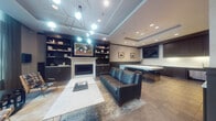 Located at 1115 SW Market St in Portland, Oregon, this refined building lobby offers a polished and welcoming first impression. The entry corridor sets an elevated tone with gleaming marble-look tile flooring, coffered ceilings with recessed and cove lighting, deep navy wainscoting, and a distinctive geometric patterned accent wall. A framed artwork serves as a striking focal point at the end of the hall, while glass entry doors allow natural light to filter through from the exterior.

Beyond the entry, the lounge area exudes sophisticated comfort. A built-in fireplace is framed by floor-to-ceiling dark cabinetry and open shelving, curated with books and decorative objects. A marble-topped coffee table anchors the seating arrangement, which includes a deep teal leather sofa and mid-century modern accent chairs with plaid upholstery. A sculptural multi-globe chandelier overhead adds a contemporary artistic touch.

Large street-facing windows flood the lounge with natural daylight and offer views of the tree-lined neighborhood outside. Adjacent to the lounge is a well-appointed kitchenette featuring dark cabinetry, a white stone countertop island with bar seating, a wall-mounted television, and under-counter appliances — ideal for resident gatherings and events.