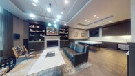 Located at 1115 SW Market St in Portland, Oregon, this refined building lobby offers a polished and welcoming first impression. The entry corridor sets an elevated tone with gleaming marble-look tile flooring, coffered ceilings with recessed and cove lighting, deep navy wainscoting, and a distinctive geometric patterned accent wall. A framed artwork serves as a striking focal point at the end of the hall, while glass entry doors allow natural light to filter through from the exterior.

Beyond the entry, the lounge area exudes sophisticated comfort. A built-in fireplace is framed by floor-to-ceiling dark cabinetry and open shelving, curated with books and decorative objects. A marble-topped coffee table anchors the seating arrangement, which includes a deep teal leather sofa and mid-century modern accent chairs with plaid upholstery. A sculptural multi-globe chandelier overhead adds a contemporary artistic touch.

Large street-facing windows flood the lounge with natural daylight and offer views of the tree-lined neighborhood outside. Adjacent to the lounge is a well-appointed kitchenette featuring dark cabinetry, a white stone countertop island with bar seating, a wall-mounted television, and under-counter appliances — ideal for resident gatherings and events.