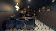 Located at 375 Camino De La Reina in San Diego, CA, this single-floor lounge and bar venue delivers a striking, moody aesthetic throughout its thoughtfully curated interior. Deep charcoal and teal walls set the tone, while recessed lighting and warm brass wall sconces cast an intimate glow across the space.

The main lounge area is anchored by a bold red velvet banquette sofa paired with zebra-print accent chairs and vibrant teal side tables, creating an eclectic, high-energy atmosphere. A gallery of distinctive original artwork adorns the walls, including a standout mixed-media piece alongside sculptural wall installations that reinforce the venue's artistic identity.

The bar area is a design centerpiece, featuring dark marble countertops, gold-accented pendant lighting with globe fixtures, and a well-stocked back bar with open shelving. Tufted navy velvet bar chairs with brass legs provide refined seating at the bar counter, and draft beer taps and full bar equipment speak to its functional sophistication.

A dedicated screening room rounds out the space, equipped with a large wall-mounted display and furnished with the same plush zebra-print lounge chairs and teal accent tables found throughout, ensuring a cohesive aesthetic experience. The geometric black-and-white patterned tile floor flows consistently through every area, tying the entire venue together with graphic elegance.
