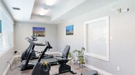 This dedicated fitness room is designed to support a well-rounded workout routine within a compact, single-level layout. The space is finished with light gray walls, white baseboards, and durable flooring that transitions between carpet and polished concrete, giving the room a clean and functional aesthetic. A tray ceiling adds a subtle architectural detail overhead, while flush-mounted fluorescent lighting ensures the space is well-lit throughout.

Large windows fitted with horizontal blinds line one wall, allowing natural light to filter in and offering views of the surrounding urban environment. The cardio equipment selection includes a treadmill, an elliptical trainer, and a recumbent exercise bike, providing users with diverse options for aerobic exercise. A cable machine with a weight stack offers strength training capabilities, and a rowing-style machine rounds out the fitness offerings.

Decorative touches such as framed cityscape and mountain landscape prints add visual interest to the walls, while potted plants stationed near the windows bring a refreshing natural element to the environment. A hand sanitizer dispenser and supply shelving near the entrance reflect thoughtful attention to cleanliness and user convenience. A water station is also present, ensuring hydration needs are met during workouts. Overall, the fitness room delivers a practical, organized, and inviting exercise environment.