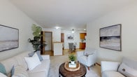 This single-floor space presents an open-concept layout that seamlessly connects the living area, dining space, and kitchen, creating a cohesive and functional environment. The living area is well-proportioned, accommodating a comfortable seating arrangement centered around a round wooden coffee table atop a neutral area rug. White walls and light-toned flooring contribute to a bright, airy atmosphere throughout. A glimpse into an adjacent room reveals a closet with built-in shelving, adding practical storage options. The dining area, positioned centrally within the open layout, is lit by a ceiling fan with an integrated light fixture and is furnished with upholstered chairs. The kitchen, visible in the background, features warm wood cabinetry, light countertops, and standard appliances, offering a classic and efficient workspace. A peninsula or breakfast bar serves as a subtle divider between the kitchen and the living and dining zones. Decorative touches, including a large potted plant and framed wall art, enhance the welcoming ambiance of the space.