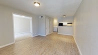This single-floor space offers a clean, modern layout designed for comfortable everyday living. The main living area is finished with light hardwood-style flooring that extends throughout, creating a warm and cohesive aesthetic. Neutral gray walls and flush ceiling lighting keep the space feeling bright and airy.

The open-concept design connects the living area directly to the kitchen, which features a raised bar-style counter that serves as both a functional workspace and a casual dining or serving surface. The kitchen is equipped with overhead cabinetry finished in white, providing ample storage, along with a built-in microwave and additional appliances. The white cabinetry contrasts tastefully with the neutral tones of the surrounding walls.

A dedicated in-unit laundry area is conveniently tucked within the main living space, housing a washer and dryer side by side — a practical feature that adds significant day-to-day convenience. A doorway from the main area leads to an adjacent room, carpeted in a light neutral tone, offering a comfortable and quiet retreat.

The space also includes a closeted utility or storage area, visible HVAC venting for climate control, and clean white baseboards throughout that lend a polished, well-maintained appearance. Overall, the layout is functional, thoughtfully arranged, and move-in ready.