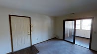 This single-floor residential space offers a straightforward and functional layout designed for comfortable everyday living. The main living area features wall-to-wall carpeting in a neutral tone, providing a warm and soft underfoot feel, while a contrasting dark hardwood floor section near the entryway adds a distinct visual transition. Dark brown wood trim frames the doorways and baseboards throughout, lending a classic, cohesive look to the interior. The front entry door, finished in white with brass hardware, opens directly into the living space, keeping the layout open and accessible. A sliding glass door with a dark frame is a standout feature, allowing natural light to filter into the room and providing a view and access to an adjacent area, likely a patio or exterior space. The walls are painted in a clean, neutral white, creating a bright and versatile backdrop suited to a variety of décor styles. A floor-level vent indicates a central air and heating system serviced throughout the unit. The overall ambiance is clean, understated, and move-in ready, offering a blank canvas for personalization.