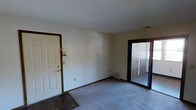 This single-floor residential space offers a straightforward and functional layout designed for comfortable everyday living. The main living area features wall-to-wall carpeting in a neutral tone, providing a warm and soft underfoot feel, while a contrasting dark hardwood floor section near the entryway adds a distinct visual transition. Dark brown wood trim frames the doorways and baseboards throughout, lending a classic, cohesive look to the interior. The front entry door, finished in white with brass hardware, opens directly into the living space, keeping the layout open and accessible. A sliding glass door with a dark frame is a standout feature, allowing natural light to filter into the room and providing a view and access to an adjacent area, likely a patio or exterior space. The walls are painted in a clean, neutral white, creating a bright and versatile backdrop suited to a variety of décor styles. A floor-level vent indicates a central air and heating system serviced throughout the unit. The overall ambiance is clean, understated, and move-in ready, offering a blank canvas for personalization.