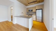 Located at 5630 Rue De Marseille in Montréal's east end, this one-bedroom, one-bathroom unit showcases a contemporary industrial-modern aesthetic throughout. The open-concept living area is defined by striking exposed concrete ceilings that contrast beautifully with crisp white walls and warm light wood flooring, creating a bright and airy atmosphere. A large window with balcony access floods the living space with natural light and offers views of surrounding greenery.

The kitchen is thoughtfully designed with an L-shaped layout and a peninsula island topped with a sleek grey countertop and an undermount sink. Light wood cabinetry, a tiled backsplash, and a full suite of stainless steel appliances — including a refrigerator, range, and built-in microwave — complete the functional and stylish cooking space. Track lighting overhead adds a modern touch.

The bedroom is a calm and comfortable retreat, featuring the same exposed concrete ceiling, a dedicated closet with sliding doors, a window with natural views, and a baseboard heating unit. A feature concrete accent wall adds industrial character to the room.

The bathroom is clean and functional, finished with large-format ceramic tile flooring, a glass-enclosed shower, and accessibility-friendly grab bars. A hallway connects all rooms, ensuring a practical and well-organized layout throughout this efficient urban home.