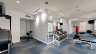 Located in New Orleans, LA, this single-floor fitness center is thoughtfully designed to support a wide variety of workout routines. The space is finished with a patterned blue and gray modular carpet that adds visual interest while providing a functional, comfortable surface underfoot. Clean white walls and a combination of recessed and track lighting create a bright, energizing atmosphere throughout.

The main workout area is anchored by a full dumbbell rack lined against a large floor-to-ceiling mirror, which visually expands the space and aids in form correction during training. A freestanding red punching bag serves as a central focal point, complemented by a flat bench for strength training exercises. A StairMaster step mill is positioned near the glass-paneled entrance, making efficient use of the transitional zone. Additional equipment includes a rowing machine, a treadmill, resistance training stations, and a Pilates reformer, offering a comprehensive range of cardio and strength options.

Natural light filters in through multiple windows, with views of the surrounding exterior, lending the space an open and welcoming feel. A dedicated section features kettlebells and accessories for functional fitness training. Folding chairs along one wall provide a convenient rest area. The glass-door entryway ensures easy access and maintains visual connectivity throughout the facility.