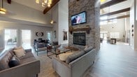 Located in Lebanon, Tennessee, this single-floor space presents an impressive open-concept layout defined by dramatic soaring ceilings and exposed wooden beams that lend a warm, rustic-modern character throughout. The expansive main living area serves as the centerpiece of the space, anchored by a floor-to-ceiling stacked stone fireplace with a wood mantel and mounted flat-screen television above. Multiple seating arrangements are thoughtfully positioned throughout the open area, creating distinct yet connected zones ideal for gathering and socializing. Pendant lighting and a statement chandelier complement the natural light streaming in through multiple windows, enhancing the airy, welcoming atmosphere. Wide-plank hardwood flooring flows seamlessly across the entire space, unifying each area cohesively. The kitchen features rich dark wood cabinetry, a classic subway tile backsplash, stainless steel appliances, and a large granite-topped island with a reclaimed wood base, accompanied by industrial-style bar stools. A billiards table is also present, adding a recreational dimension to the layout. The overall ambiance blends contemporary design with rustic touches, creating a versatile and inviting environment.