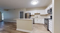 This single-floor residence offers a clean, modern aesthetic defined by an open-concept layout that seamlessly connects the kitchen and living area. The kitchen is a standout feature, equipped with crisp white shaker-style cabinetry, light-toned countertops, and a full suite of stainless steel appliances including a refrigerator, range with over-the-range microwave, and dishwasher. A herringbone-patterned tile backsplash adds a refined decorative touch, while a central island provides additional prep space and a natural visual anchor for the room. Wood-style plank flooring flows consistently throughout the main living areas and bedrooms, contributing warmth and a cohesive feel to the interior. The living area is generously proportioned and benefits from natural light streaming in through large windows that frame views of mature trees outside. The bedrooms are well-sized, each featuring the same wood-style flooring and windows that bring in ample daylight. The bathroom is finished with large-format marble-look tile flooring, a white vanity with a mirror, and brushed nickel hardware accents. Neutral warm-beige wall tones throughout the space create a calm, inviting atmosphere, while textured ceilings and clean white trim complete the polished, move-in-ready presentation.