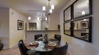 This well-appointed single-floor residence showcases a thoughtfully designed interior that blends elegance with everyday functionality. The dining area serves as a focal point of the space, centered around a round glass-top dining table surrounded by sleek dark leather chairs, creating an intimate setting ideal for entertaining. A contemporary chandelier with cylindrical frosted shades hangs overhead, casting warm, inviting light across the room, complemented by track lighting that adds depth and ambiance. Crown molding lines the ceiling throughout, adding a refined architectural detail that elevates the overall aesthetic. A decorative arrangement of dark-framed rectangular mirrors adorns one wall, visually expanding the space and adding a sophisticated design element. The dining area flows seamlessly into the kitchen, which features a granite or stone-topped breakfast bar that doubles as a pass-through, allowing for easy interaction between the two spaces. The kitchen is further illuminated by pendant lighting, enhancing its open and accessible feel. Neutral warm tones on the walls and light hardwood flooring unify the space, creating a cohesive and welcoming atmosphere throughout. The overall design strikes a balance between modern style and comfortable, livable elegance.