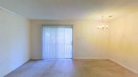 This single-floor space offers a clean, functional layout designed for comfortable everyday living. The main living area is a standout feature, characterized by its open feel and abundant natural light streaming through a full-width sliding glass door dressed with vertical blinds. The sliding door provides both a visual connection to the outdoors and convenient exterior access. Overhead, an elegant multi-arm chandelier adds a refined decorative touch to the space, lending a sense of classic style to the room. The flooring throughout is fitted with soft, neutral-toned carpeting that contributes to a warm and inviting atmosphere. The walls are finished in a light, neutral tone that complements the brightness of the space and enhances its airy quality. Clean white baseboards frame the room neatly, adding a crisp, finished look to the overall aesthetic. The combination of natural light, thoughtful lighting fixtures, and understated finishes creates a welcoming ambiance throughout. The layout is well-suited for versatile furniture arrangements, offering flexibility for both relaxed living and functional use of the available space.
