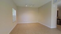 This two-floor space presents a clean, minimalist aesthetic defined by neutral tones and practical finishes. The pictured room showcases warm beige walls complemented by white baseboards, creating a bright and open atmosphere. Light tan ceramic tile flooring extends throughout, offering durability and ease of maintenance. A track lighting fixture with multiple adjustable heads is mounted to the ceiling, providing flexible and even illumination across the room. The ceiling height lends a comfortable sense of openness to the space. A window fitted with horizontal blinds allows natural light to filter in, brightening the room while offering privacy control. A ceiling-mounted HVAC vent indicates a central air system serving the space. The walls feature standard electrical outlets positioned conveniently throughout. The overall interior palette—combining beige walls, light tile, and white trim—creates a cohesive, neutral canvas well-suited to a variety of uses and furnishing styles. The two-floor layout suggests additional living or functional space beyond what is pictured, offering versatility in how the property can be utilized.
