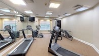 This fitness room is a comprehensively outfitted exercise facility designed to support a full range of workout routines. The space is finished with neutral beige walls and wall-to-wall carpet flooring, creating a clean and comfortable environment for physical activity. Recessed flush-panel ceiling lights provide consistent, bright illumination throughout the room, supplemented by natural light from a window that offers a view of a green outdoor setting.

The facility is equipped with multiple cardio machines, including treadmills and an elliptical trainer, catering to a variety of cardiovascular exercise preferences. A multi-station cable strength-training machine anchors one side of the room, accompanied by an adjustable bench and instructional exercise charts mounted nearby. A recumbent stationary bike rounds out the equipment selection, offering lower-impact workout options. Wall-mounted flat-screen televisions provide entertainment during exercise sessions.

A full wall of floor-to-ceiling mirrors visually expands the space and allows users to monitor their form during workouts. Organizational elements such as a wall-mounted information and supply station, clipboards, and a wall clock contribute to the functional layout. Double doors provide easy access to and from the facility. The overall design is practical and well-maintained, making this fitness room a convenient and versatile amenity.