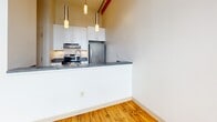 Located at 100–144 Clifford St in Providence, Rhode Island, this single-floor unit reflects the character of a converted industrial building, blending historic architectural details with modern updates throughout. Original hardwood floors run continuously across the main living areas, lending warmth and a sense of continuity to the space.

The open-concept kitchen features white cabinetry, stainless steel appliances including a refrigerator and electric range, dark countertops, and a peninsula that separates it from the living area. Pendant lighting hangs above the peninsula, adding a contemporary touch. Exposed structural beams are visible overhead, nodding to the building's industrial past.

A hallway connects the main living area to additional rooms, with elevated ceilings contributing to an airy, loft-like atmosphere. One room showcases a striking exposed brick wall paired with large, black-framed industrial windows that frame views of the surrounding Providence cityscape and flood the space with natural light.

The bathroom is finished with light tile flooring, a full tub and shower surround clad in classic white subway tile, and accessibility grab bars. Throughout the unit, the combination of raw historic materials and clean modern finishes creates a distinctive, urban aesthetic.