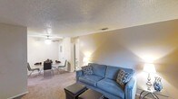 This single-story space features an open-concept layout that seamlessly connects the living and dining areas, creating a cohesive and functional environment. The living area is anchored by a blue upholstered sofa accented with patterned throw pillows, paired with a dark ottoman that serves as a coffee table. Neutral-toned carpet flows throughout the space, adding warmth underfoot.

A ceiling fan with a light fixture is centrally positioned overhead, providing both ventilation and ambient lighting. The textured popcorn ceiling adds a classic residential character to the interior. Natural transitions between zones are defined by furniture placement rather than physical walls, maintaining an airy, open feel.

The dining area is situated adjacent to the living space and features a glass-top table surrounded by contemporary chairs, suitable for everyday meals or casual gatherings. A floor lamp near the dining zone provides warm, localized lighting. Doors visible in the background suggest access to additional rooms or storage. The overall ambiance is practical and understated, with a neutral color palette that lends itself well to personalization.