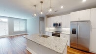 Located at 2815 Capital Blvd in Raleigh, NC, this modern single-floor residence offers a thoughtfully designed open-concept layout with clean finishes and contemporary style throughout. The kitchen serves as the heart of the home, showcasing a large central island topped with speckled granite countertops, an undermount sink, and striking geometric pendant lights. Crisp white shaker cabinetry, a decorative tile backsplash, and a full suite of stainless steel appliances — including a French door refrigerator, electric range, microwave, and dishwasher — complete the space. Warm-toned hardwood flooring flows seamlessly from the kitchen into the open living area, which is bathed in natural light from multiple windows and a glass-panel entry door. A built-in mudroom nook with shelving and hooks sits conveniently near the entry. The primary bathroom features a glass-enclosed walk-in shower alongside direct access to a spacious walk-in closet outfitted with multi-level wire shelving and carpeted flooring. A secondary walk-in closet offers additional storage with the same practical wire shelving system. A half bath with a window provides added convenience. Neutral gray walls and recessed lighting throughout create a bright, cohesive ambiance across the entire home.
