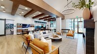 This expansive single-floor community clubhouse delivers a sophisticated blend of modern farmhouse and contemporary design across a generously proportioned open layout. Soaring ceilings define the main living and lounge area, where rich, dark wood exposed beams provide striking architectural contrast against crisp white walls. Wide-plank light wood flooring flows throughout, anchoring the space with warmth and continuity.

The lounge zone is anchored by plush seating arrangements, including neutral sectional sofas and cognac leather accent chairs, centered around a sleek fireplace with a mounted flat-screen TV above. Statement ring chandeliers illuminate each zone, adding a refined decorative element throughout.

A dedicated dining area features a large communal table with Windsor-style seating, ideal for group gatherings. Adjacent to this, a service bar area showcases bold geometric patterned tile flooring, a dark-topped island with integrated sink, and floor-to-ceiling black-framed glass walls that connect to an outdoor terrace, flooding the space with natural light and garden views.

A more intimate lounge nook features decorative wallpaper, wainscoting, built-in bench seating, and a billiards table, offering a cozy retreat within the larger space. Black-framed steel-and-glass partitions elegantly define zones while preserving the open, airy atmosphere throughout.