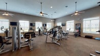 This fitness center offers a comprehensive workout environment designed to accommodate a wide range of exercise routines. The space is generously sized, with neutral-toned walls, carpet tile flooring, and multiple ceiling fans that contribute to a comfortable atmosphere throughout.

The main workout area is lined with large windows fitted with white blinds, allowing ample natural light to fill the room while offering views of the surrounding exterior. Multiple wall-mounted flat-screen televisions are positioned throughout, providing entertainment during cardio sessions.

Cardio equipment is well-represented, with several treadmills, elliptical machines, and stationary bikes arranged to maximize floor space. A dedicated strength training section houses a variety of cable and resistance machines suitable for full-body workouts. A separate free weights area features a complete rack of dumbbells, kettlebells, and medicine balls alongside a adjustable bench for targeted training.

A mirrored wall section anchors a functional fitness zone stocked with stability balls, balance discs, and step platforms, ideal for flexibility and core work. A large wall-mounted television in this area supports guided workout sessions. The facility is accessed through glass double doors and includes clearly posted guidelines, ensuring a well-organized and user-friendly experience.
