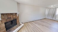 This two-storey, 2-bedroom, 1-bathroom unit in Calgary, Alberta offers a well-organized split-level layout across two floors. The main floor houses the living room, which serves as the focal point of the home with its striking stone-surround wood-burning fireplace, light hardwood flooring, and a textured ceiling. A large front-facing window fills the space with natural light and provides a view of the surrounding area, while a front entry door opens directly into this welcoming living area. A staircase with a metal railing leads to the upper floor, which contains both bedrooms, a full bathroom, and connecting hallways. The two bedrooms feature the same light hardwood flooring, neutral white walls, and individual windows fitted with horizontal blinds, allowing for adjustable natural light. Baseboard heating is present throughout the unit. The bathroom is equipped with a full-size soaking tub and shower combination, accented by large-format beige tile surround, alongside a vanity with a white countertop and a mirrored medicine cabinet. The clean, neutral palette and consistent flooring throughout both floors contribute to a cohesive and functional living environment.