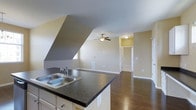 This single-floor space showcases an open-concept layout that seamlessly connects the kitchen and living areas. The kitchen features a peninsula with dark granite-style countertops and a double stainless steel sink, complemented by white cabinetry beneath. The flooring throughout is rich, dark hardwood, adding warmth and continuity across the space.

A notable architectural feature is the angled vaulted ceiling, which adds visual interest and a sense of elevated volume to the space. The living area is equipped with a ceiling fan with a light fixture, providing both airflow and ambient lighting. Neutral beige and gray wall tones create a clean, versatile backdrop throughout.

Natural light enters through strategically placed windows, while additional lighting fixtures ensure the space remains well-lit. A hallway visible from the main living area leads to additional rooms, suggesting a functional and well-organized floor plan. The open layout, combined with the architectural ceiling detail and quality finishes, gives the space a practical yet polished character.