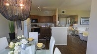 This single-floor residence offers an open-concept design that seamlessly connects the kitchen, dining, and living areas, creating a cohesive and functional living environment. The kitchen is outfitted with warm-toned wooden cabinetry, black appliances including a built-in microwave and gas range, a full-size refrigerator, and a breakfast bar with a white countertop that serves as both a prep space and casual dining area. The cabinetry provides generous storage, and the layout ensures an efficient workflow throughout the space. Adjacent to the kitchen, the dining area is anchored by a glass-top table surrounded by sleek white chairs, accented by an eye-catching beaded chandelier overhead that adds a touch of elegance. Rich, dark hardwood-style flooring runs throughout the main living areas, lending warmth and continuity to the space. Track lighting along the ceiling provides versatile illumination, while high ceilings in the kitchen zone enhance the sense of openness. A glimpse into the background reveals an additional living or lounge area, suggesting a generous overall footprint. The combination of modern finishes, warm wood tones, and thoughtful design details gives this residence a welcoming yet sophisticated ambiance.