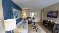Located at 9015 Ten Mile Rd in Knoxville, Tennessee, this single-floor residence offers a thoughtfully designed living space that blends comfort with contemporary style. The living room serves as the centerpiece of the home, featuring a striking deep blue accent wall adorned with a gallery of framed abstract artwork, creating a bold visual focal point. Neutral-toned walls throughout complement the vibrant accents, maintaining a balanced and welcoming atmosphere. Light wood-style flooring runs throughout, adding warmth and continuity to the space. The living area is well-appointed with a spacious sectional sofa dressed in an array of patterned throw pillows in coordinating blues and yellows, alongside a classic wingback accent chair. A glass-top coffee table with a sculptural base anchors the seating arrangement atop a layered area rug. A wall-mounted mini-split unit provides efficient climate control, while a window allows natural light to filter into the room. A dark wood sideboard adds functional storage and a rich contrast to the lighter palette. A hallway visible from the living area connects to additional rooms, suggesting a practical and well-organized floor plan throughout this inviting Knoxville home.