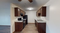 This single-floor space offers a thoughtfully designed interior with a focus on functionality and classic style. The galley-style kitchen serves as a central highlight, featuring rich, dark wood cabinetry that lines both walls, providing generous storage and a warm, inviting aesthetic. Light granite-look countertops contrast beautifully against the dark cabinetry, creating a visually balanced workspace. The kitchen is equipped with black appliances, including a gas range, overhead range hood, and a full-sized refrigerator, making it well-suited for everyday cooking. A stainless steel sink is positioned on the opposing counter wall, complemented by additional upper and lower cabinet storage. The kitchen floor features wood-look plank flooring in a warm tone, adding to the overall cohesive design. A ceiling fan is mounted overhead, providing both air circulation and ambient lighting, while a flush-mount ceiling light fixture further brightens the space. The textured ceiling adds a subtle architectural detail throughout. The overall ambiance of the space is cozy and practical, combining durable finishes with a classic design that offers both comfort and utility.