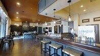 This single-floor social space is thoughtfully designed to blend modern sophistication with rustic warmth, creating an inviting atmosphere ideal for gathering and entertaining. At the heart of the space is a long, rich wood-topped bar lined with industrial-style barstools featuring reclaimed wood seats, offering ample seating for guests. Above the bar, a row of clear glass pendant lights hangs from the ceiling, casting a warm, ambient glow throughout the area. The bar itself is well-appointed with a built-in sink, storage cabinetry in a muted gray-blue finish, and overhead cabinets that extend toward the ceiling, maximizing vertical space. Beyond the bar, the lounge area invites relaxation with comfortable seating arranged around a striking brick fireplace, which serves as a focal point and adds a cozy, residential character to the space. The ceilings throughout are elevated, with a warm orange-toned accent ceiling above the bar zone that transitions into lighter tones in the lounge area, adding visual interest and depth. Recessed lighting, decorative pendants, and a globe light fixture layer the illumination throughout. The polished concrete floors tie the design together, offering a clean, durable, and contemporary foundation that complements the overall modern-rustic aesthetic.