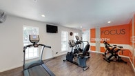 The fitness center is a bright, single-level space designed to encourage an active and healthy routine. The room features hardwood-style flooring throughout, providing a clean and durable surface well-suited for exercise equipment and movement. White walls keep the space feeling open and airy, while recessed lighting and multiple windows along one wall allow natural light to fill the room, creating an inviting workout environment.

The space is equipped with a selection of cardio machines, including a treadmill, elliptical, and stationary bike, offering residents a range of options for cardiovascular exercise. A wall-mounted flat-screen television is positioned near the windows, adding entertainment value during workouts. A decorative clock is also visible on the wall, adding a functional touch.

A standout feature of the fitness center is the bold orange and white accent wall, which displays motivational text referencing focus and strength. This graphic element adds energy and visual interest to the space, reinforcing its purpose as a place dedicated to fitness and wellness. The overall design is clean, functional, and modern.