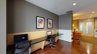 This single-floor space is designed as a functional and stylish business center, offering a well-appointed environment for focused work. The workstation area features a sleek, built-in desk surface with warm wood-toned cabinetry below, providing ample workspace for multiple users simultaneously. Ergonomic task chairs complement the practical setup, while recessed lighting overhead ensures a well-lit environment throughout the day. The walls are finished in a contemporary cool gray, accented by a two-tone treatment with a lighter lower panel, lending the space a polished, professional feel. Two bold, colorful framed artworks adorn the wall above the workstations, adding a vibrant artistic touch to the otherwise understated palette. Rich, dark hardwood flooring runs throughout, grounding the space with warmth and elegance. An open passage connects the workstation area to an adjacent lounge or common area, which is visible in the background and features warm amber-toned walls and casual seating, creating a sense of openness and flow between functional and relaxed zones. Overall, the space balances productivity and comfort through thoughtful design, quality finishes, and a cohesive modern aesthetic.