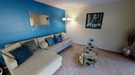 This single-floor residence makes a memorable first impression with its thoughtfully designed living area, where a bold cobalt blue accent wall immediately draws the eye and sets a vibrant, contemporary tone. The living room is centered around a spacious sectional sofa dressed in coordinating blue and neutral throw pillows, creating an inviting and cohesive look. A sleek, circular glass coffee table anchors the seating area, reflecting light and adding a touch of modern elegance to the space. Soft, neutral carpeting runs throughout, providing warmth and comfort underfoot, while crisp white walls on the surrounding surfaces offer a clean contrast to the vivid accent wall. A stylish floor lamp provides warm ambient lighting, complementing the decorative framed wall art that adds a splash of color and artistic character to the room. A geometric-framed mirror mounted on the accent wall further enhances the sense of depth and dimension in the space. The overall ambiance is modern and lively, blending bold color choices with contemporary furnishings to create a living environment that feels both energetic and comfortable — ideal for relaxing or entertaining.