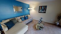 This single-floor residence makes a memorable first impression with its thoughtfully designed living area, where a bold cobalt blue accent wall immediately draws the eye and sets a vibrant, contemporary tone. The living room is centered around a spacious sectional sofa dressed in coordinating blue and neutral throw pillows, creating an inviting and cohesive look. A sleek, circular glass coffee table anchors the seating area, reflecting light and adding a touch of modern elegance to the space. Soft, neutral carpeting runs throughout, providing warmth and comfort underfoot, while crisp white walls on the surrounding surfaces offer a clean contrast to the vivid accent wall. A stylish floor lamp provides warm ambient lighting, complementing the decorative framed wall art that adds a splash of color and artistic character to the room. A geometric-framed mirror mounted on the accent wall further enhances the sense of depth and dimension in the space. The overall ambiance is modern and lively, blending bold color choices with contemporary furnishings to create a living environment that feels both energetic and comfortable — ideal for relaxing or entertaining.