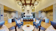 This single-floor leasing office is designed with a refined, hospitality-inspired aesthetic that immediately sets a welcoming tone. The centerpiece of the space is a grand lounge area distinguished by soaring coffered ceilings accented with rich dark wood beams. Three dramatic globe chandeliers with crystalline detailing hang overhead, casting a warm, inviting glow across the light hardwood floors below. A symmetrical seating arrangement features plush blue wingback chairs and a coordinating sofa, centered around a sleek white round coffee table, creating an approachable yet upscale atmosphere. A decorative fireplace flanked by tall tropical plants and framed artwork anchors the far wall, complemented by a geometric-front credenza that adds a bold design statement. Floor-to-ceiling glass storefront windows at the entrance offer clear views to the surrounding streetscape, flooding the space with natural light. A dedicated leasing desk area sits to one side, framed by an ornate patterned partition screen, providing a semi-private workspace. A well-appointed beverage station featuring marble countertops, mirrored tile backsplash, and built-in glass-door beverage coolers offers a polished amenity for visitors. Throughout, the design balances modern sophistication with warm, residential comfort.