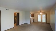 This single-floor space offers an open and functional layout connecting the living area and kitchen. The living room is finished with wall-to-wall carpet, a textured ceiling, and a ceiling fan that enhances air circulation while adding a subtle decorative touch. Dark baseboards provide a clean contrast against the neutral-toned walls throughout.

The kitchen is positioned toward the rear of the main living area and features warm-toned wooden cabinetry, a white countertop, and white appliances, including a dishwasher. The kitchen's open sightlines into the main living space support an easy flow between cooking and entertaining areas.

A standout feature of the space is the set of French doors in the living area, framed with divided-light glass panels that invite a generous amount of natural light into the interior, creating a bright and welcoming ambiance. A doorway off the main living area leads to an additional room, visible in the background, which appears to include a washer/dryer hookup or utility area, adding practical convenience to the layout. Overall, the space presents a straightforward, functional design suited for comfortable everyday living.