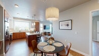 This thoughtfully designed single-floor residence offers a seamless open-concept layout that connects the kitchen, dining, and living areas into one cohesive living space. The kitchen is a standout feature, showcasing a striking two-tone cabinetry design that pairs rich warm wood lowers with crisp white uppers, complemented by quartz countertops, a subway tile backsplash, stainless steel appliances, and a generous island with bar seating. Natural light pours in through large windows fitted with blinds, brightening the warm wood-tone flooring found throughout. The dining area centers around a round table beneath a sculptural drum pendant light, while a nearby entryway features a decorative console and round mirror. The living room is highlighted by a bold accent wall with patterned wallpaper, a ceiling fan, and sliding glass doors that open to an outdoor space. The bedroom continues the warm aesthetic with a rich terracotta accent wall and ceiling fan. Two bathrooms are finished with warm wood vanities and clean white quartz countertops — a full bath featuring a double-sink vanity with ample storage, and a sleek powder-style bath. The overall ambiance is modern yet warm, with a cohesive mid-century-inspired design sensibility throughout.