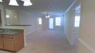 This single-floor space features a generously sized open living area finished with plush wall-to-wall carpeting in a neutral tone, creating a warm and inviting atmosphere. The ceiling draws immediate attention with its distinctive patterned texture, complemented by elegant crown molding that runs along the perimeter of the room, adding a refined architectural detail. A ceiling fan with a light fixture is centrally mounted, providing both air circulation and ambient lighting.

Natural light fills the space through multiple windows, including a pair of sidelights flanking a glass-paneled door that leads to an outdoor area, offering views and additional brightness. A secondary window is visible in an adjacent room, further enhancing the light-filled quality of the interior.

To one side, a partial view of the kitchen reveals a raised countertop with a dark granite or stone surface, supported by cabinetry in a warm wood finish, suggesting an open-concept layout that connects the kitchen to the living area. The overall design balances functional and aesthetic elements, with clean white walls and classic trim work contributing to a timeless, well-maintained interior.