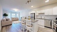 Located at 67-71 Minerva Street in Derby, CT, this well-appointed 1-bedroom, 1-bathroom residence offers a thoughtfully designed single-floor layout finished with consistent light hardwood flooring throughout. The open-concept kitchen flows seamlessly into the living and dining areas, creating an inviting, connected space. The kitchen is equipped with crisp white shaker cabinetry, granite countertops, a subway tile backsplash, stainless steel appliances, and a central island with bar seating — ideal for both everyday meals and casual entertaining. The living room is generously sized, featuring a bay window arrangement that draws in abundant natural light and provides views of the surrounding area. Soft neutral tones and clean lines give the space a calm, modern aesthetic. The primary bedroom is notably spacious, benefiting from multiple windows that bathe the room in sunlight, complemented by light gray walls and hardwood-style flooring. The full bathroom is finished with a white vanity featuring granite countertops, wood-look tile flooring, a sliding glass walk-in shower, and a bright window. A connecting hallway ties the layout together efficiently. Throughout, the space maintains a bright, contemporary ambiance with neutral finishes that offer a clean and move-in-ready appeal.