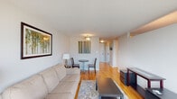 This single-floor residence offers a thoughtfully designed layout that balances comfort and functionality. The living area serves as the heart of the space, featuring warm hardwood flooring, neutral walls, and an abundance of natural light streaming through large floor-to-ceiling windows that frame sweeping panoramic views of the surrounding cityscape and greenery. The open feel is complemented by tasteful wall art and a cozy seating arrangement centered around a coffee table and area rug, with a dedicated workspace tucked neatly by the window. A compact dining nook with a bistro-style table and chairs provides an intimate setting adjacent to the main living zone. The kitchen is efficiently designed, showcasing gray cabinetry, dark granite countertops, a stainless steel gas range with an over-the-range microwave, and a stainless steel sink — all set atop continuous hardwood floors. The bathroom is clean and functional, featuring white tile walls, a tub-shower combination, a warm wood vanity with a wide mirror framed by globe lighting, and classic mosaic tile flooring. Together, these spaces create a cohesive, light-filled residence with a comfortable and practical ambiance throughout.