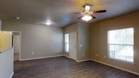 This single-floor space presents a straightforward and functional layout designed for comfortable everyday living. The main living area is finished with rich, dark hardwood-style flooring that contrasts attractively against the warm, neutral beige walls and white trim baseboards. The room is equipped with a classic five-blade ceiling fan with an integrated light fixture, providing both airflow and ambient lighting throughout the space. A second flush-mount ceiling light near the entryway ensures consistent illumination across the entire area. Natural light filters into the room through windows flanking the front entry door, brightening the interior and creating an open, airy feel. The front door is a traditional white panel design, providing a clean and welcoming entry point. Electrical outlets are conveniently positioned along the walls, and a ceiling-mounted HVAC vent contributes to efficient climate control. The overall design is clean and neutral, offering a versatile canvas well-suited to a variety of furnishing styles and personal preferences. The open layout of the main room allows for flexible furniture arrangement, making the space both practical and adaptable.