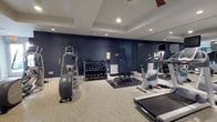 This fitness center offers a comprehensive workout environment designed to meet a variety of exercise needs. The space is defined by its striking navy blue accent walls, which contrast cleanly against white trim and a tray ceiling outfitted with recessed lighting and a ceiling fan, ensuring the room remains well-lit and comfortable during workouts. The flooring features a durable speckled terrazzo finish, both practical and visually appealing.

The cardio section is well-appointed, featuring a treadmill, an elliptical trainer, and a stationary bike, each equipped with integrated display monitors for tracking workout metrics. A dedicated free weights area includes a multi-tier dumbbell rack stocked with a full range of weights, accompanied by a flat workout bench and an exercise mat, providing ample space for strength training and stretching routines. A large wall-mounted mirror expands the sense of space and allows users to monitor their form during exercises.

Ventilation is supported by visible HVAC vents, and a wall-mounted television adds an entertainment option for users during longer sessions. The single-floor layout is efficiently organized, making the most of the available space while maintaining clear pathways between equipment. Overall, the fitness center presents a clean, modern, and fully functional environment suitable for a range of fitness routines.