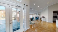 This single-floor space is defined by an open, flowing layout that seamlessly connects its key areas through polished golden tile flooring and cohesive neutral-toned walls accented with crisp white crown molding throughout.

The grand entryway immediately sets an impressive tone, featuring soaring ceilings and an abundance of natural light pouring in through floor-to-ceiling French doors on both sides. A vibrant abstract area rug anchors the foyer, offering a welcoming transition into the interior.

The main lounge area is a standout feature, furnished with teal accent chairs arranged around glass-top tables atop a boldly patterned rug. Recessed lighting illuminates the space, while a elegant tile-surround fireplace with a decorative mantle serves as the room's focal point. Large windows dressed with sheer white curtains and blinds invite ample natural light and views of the surrounding exterior.

An adjacent dining area provides a sophisticated setting with a round glass-top table and sleek modern chairs, complemented by additional seating nearby, all bathed in natural light from multiple windows.

A refreshment station tucked along one wall features a mirrored white cabinet with ornate detailing, a beverage maker, and a large wall mirror above — adding both function and style. A dedicated workstation area completes the space, offering a practical zone alongside the more social areas.