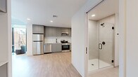 Located in Tampa's 33605 zip code, this modern studio unit at 1704 N 17th St offers a thoughtfully designed, open-concept floor plan across a single level. Light wood-tone flooring flows throughout, complementing the clean white walls and recessed lighting that keep the space feeling bright and airy.

The kitchen is a standout feature, outfitted with a full suite of stainless steel appliances — including a refrigerator, electric range, built-in microwave, and dishwasher — set against sleek grey cabinetry that extends to the ceiling for ample storage. A black matte sink fixture adds a contemporary accent. Conveniently tucked nearby is a stacked in-unit washer and dryer.

The living area is bathed in natural light through floor-to-ceiling windows that frame views of the surrounding Tampa neighborhood, creating a welcoming and versatile space. The sleeping area is integrated seamlessly into the layout, featuring light-toned walls and a wall-mounted TV.

The bathroom continues the modern aesthetic with large-format tile flooring, a frameless glass walk-in shower, a floating grey vanity with a white countertop, and matte black hardware and fixtures throughout. The overall design is cohesive, minimalist, and well-executed, making efficient use of every square foot.