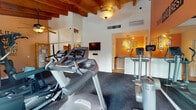 Located at 6210 N Oracle Rd in Tucson, Arizona, this single-level fitness center combines functional workout amenities with distinctive Southwestern architectural character. The space is defined by striking exposed wood beam ceilings that lend warmth and visual interest throughout, complemented by ceiling fans that enhance airflow and comfort during workouts.

The main exercise area is outfitted with a comprehensive selection of cardio equipment, including treadmills, elliptical trainers, and stair climbers, all positioned on durable rubber flooring designed to absorb impact. A dedicated strength training zone houses a multi-station cable machine, medicine balls, and resistance accessories, offering a well-rounded training experience. Wall-mounted mirrors expand the sense of space, while a mounted flat-screen television provides entertainment during workouts.

Motivational phrases are displayed prominently on the warm-toned accent walls, fostering an energizing atmosphere. Arched windows allow natural light to filter into the space, offering views of the surrounding Tucson landscape. A small adjoining area with warm lighting and framed artwork serves as a transitional lounge or refreshment nook. The overall design balances the rustic charm of traditional Southwestern aesthetics with the practical demands of a modern fitness facility.
