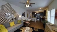 This single-floor residence showcases an open-concept design that seamlessly connects the living, dining, and kitchen areas into one cohesive, functional space. Rich hardwood-style flooring runs throughout, anchoring the warm yet contemporary aesthetic.

The living area features a ceiling fan with integrated lighting and is highlighted by a striking decorative mosaic wall accent, adding a bold artistic focal point. A comfortable sofa with vibrant accent pillows sits alongside a round coffee table, creating an inviting gathering space.

Adjacent to the living area, the dining space accommodates a bistro-style table and chairs, positioned to allow easy flow between all zones. Pendant lighting above adds a refined touch.

The kitchen is notably well-appointed, featuring dark cabinetry contrasted by stainless steel appliances, including a refrigerator and range. A breakfast bar with barstool seating provides additional casual dining options. Track lighting overhead ensures the kitchen remains bright and functional.

Overall, the space projects a modern, urban character enhanced by layered lighting, decorative details, and a cohesive color palette of grays, blacks, and gold accents.