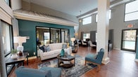 The clubhouse at District at Hurstbourne is a thoughtfully designed community space that combines modern aesthetics with functional areas for relaxing and working. The space is immediately distinguished by its dramatic double-height ceilings, which create an open, airy atmosphere throughout. Bold teal accent walls provide a striking visual anchor, complementing the warm hardwood flooring that extends across the entire space.

The main lounge area is furnished with a classic cream sofa accented by navy and mustard throw pillows, paired with mid-century modern teal accent chairs arranged around a round glass-top coffee table. A patterned area rug grounds the seating arrangement, adding texture and depth to the space. Large black-framed windows and sliding glass doors flood the interior with natural light while offering views of the surrounding community.

A dedicated workspace area features dark-toned desks and contemporary seating, providing residents a focused environment for productivity. Structural white columns add architectural interest while defining distinct zones within the open floor plan. A carpeted staircase with a rich wood banister and white balusters leads to an upper mezzanine level, further enhancing the vertical scale of the space. Overall, the clubhouse projects a polished, welcoming ambiance suited for both socializing and everyday use.