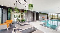 Located at 8405 Rue St-Charles in Brossard, Quebec, this contemporary gym and pool facility is designed with a clean, modern aesthetic that emphasizes natural light, open space, and thoughtful detailing throughout its single floor.

The centerpiece of the facility is the expansive indoor lap pool, enclosed by floor-to-ceiling black-framed glazing on multiple sides. This generous window wall floods the pool area with natural light and offers unobstructed views of an outdoor terrace furnished with lounge chairs, surrounded by lush landscaping. The pool deck is finished in crisp white mosaic tiling and equipped with stainless steel handrails for safe entry.

Adjacent to the pool, a dedicated relaxation zone features sleek lounge chairs arranged beneath striking circular pendant light fixtures and cascading hanging plants. A tiled open shower station and additional greenery — including wall-mounted planters — contribute to a spa-like atmosphere. Warm-toned vertical timber screens elegantly partition the pool area from the surrounding spaces.

The fitness and activity area offers a spacious, open-plan layout finished with smooth, light-toned flooring. Floor-to-ceiling windows along one wall provide garden views, while a feature wall with climbing holds and built-in seating adds a dynamic, recreational dimension. The gym area is visible with fitness equipment and full-length mirrors. Throughout the facility, abundant plantings and refined finishes create an inviting, wellness-focused environment.