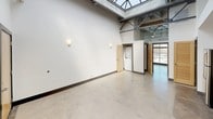 This single-floor space showcases a distinctive industrial-loft aesthetic defined by dramatically high ceilings supported by exposed steel trusses and beams. A large skylight floods the open living area with abundant natural light, highlighting the polished concrete floors that extend throughout the main space. The generous, open layout creates a versatile and airy atmosphere with clean white walls and wall-mounted sconce lighting.

The kitchen is well-appointed with dark espresso cabinetry extending to the ceiling, granite countertops, a white tile backsplash, and a full suite of stainless steel appliances including a refrigerator, range, microwave, and dishwasher. The space also includes a dedicated in-unit laundry closet housing a stacked washer and dryer, adding everyday convenience.

A louvered interior door and multiple doorways provide access to the various functional areas within the unit. The bathroom features neutral large-format floor tiles, a soaking tub with a tiled surround incorporating built-in niche shelving, brushed nickel fixtures, dark cabinetry, and a granite-topped vanity, completing the space with a refined yet understated finish.