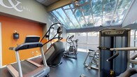 This fitness center is defined by its standout architectural feature: a expansive skylight ceiling that floods the space with natural light and offers views of the surrounding tree canopy, creating an open, airy atmosphere uncommon in typical gym environments. A ceiling fan mounted within the skylight frame aids in air circulation, enhancing comfort during workouts.

The interior walls feature a bold orange accent wall that adds energy and visual interest to the space. The flooring is finished in a dark, durable material well-suited for high-traffic fitness use. Large windows along the exterior wall further maximize natural light and provide outdoor views, reinforcing the connection to the surrounding environment.

The facility is equipped with a selection of cardio machines, including a Matrix stair climber, treadmills, and multiple elliptical trainers, catering to a range of cardiovascular workout preferences. Additional strength and fitness equipment is also present. The overall ambiance is bright, motivating, and functional, offering residents a comfortable and well-appointed space to maintain an active lifestyle.